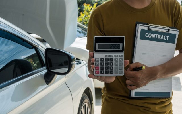 A businessman stands next to his damaged vehicle after an accide A businessman stands next to his damaged vehicle after an accident, holding a clipboard
