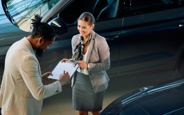 Happy black man signing contract while buying new car in auto salon. Happy African American man signing an agreement while receiving new car key from saleswoman in a showroom.
