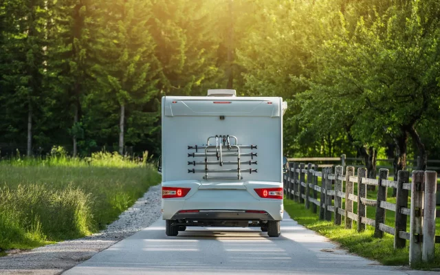 An RV travels down a tranquil dirt road bordered by a wooden fence, surrounded by tall trees and vibrant greenery. The warm glow of the setting sun illuminates the scene, creating a peaceful atmosphere that invites exploration and adventure in nature.